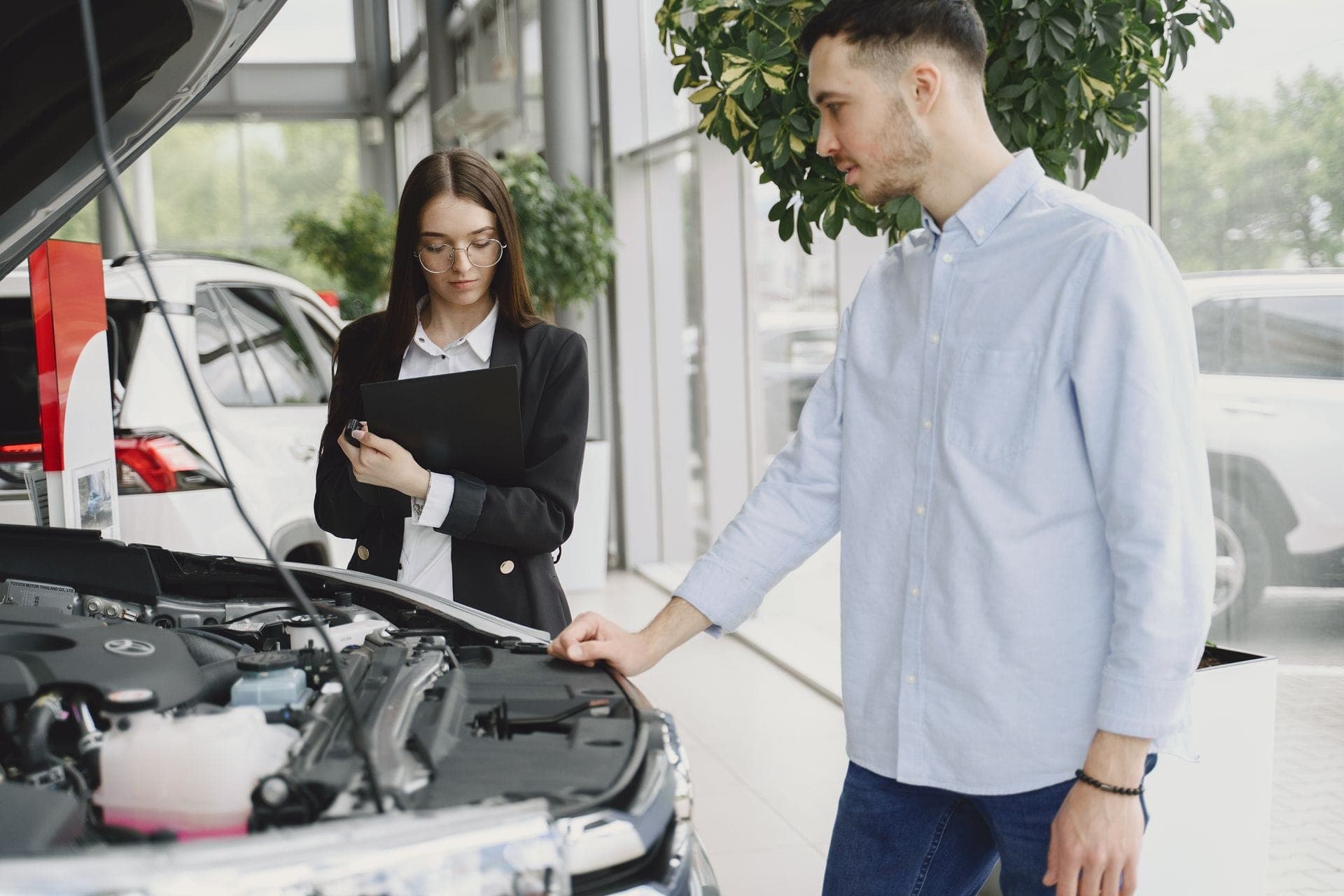 Une conseillère prend des notes sur un porte-document devant le capot ouvert d’une voiture, à côté du client